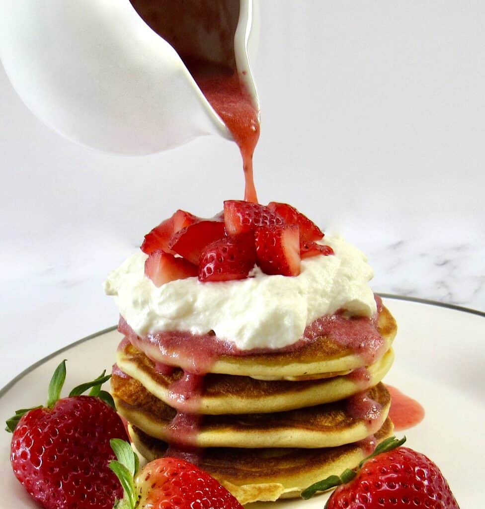 stack of pancakes with whip cream and strawberry sauce being poured over the top