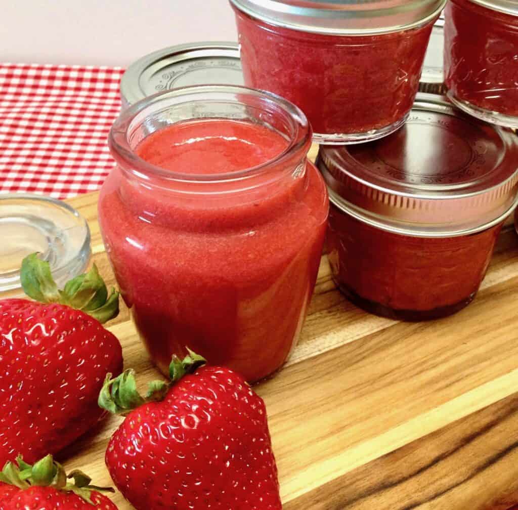 glass jar of strawberry sauce with strawberries on cutting board