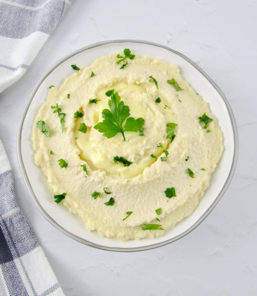 overhead view of mashed cauliflower in white bowl with butter and parsley on top