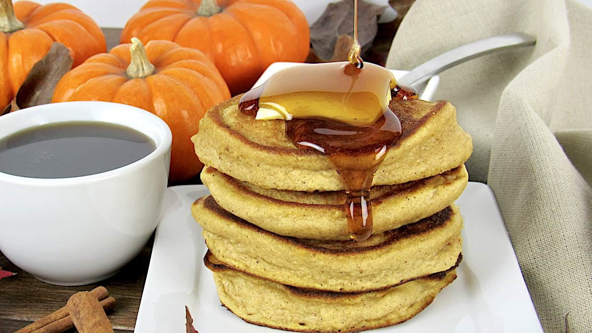 stack of Keto Pumpkin Pancakes with syrup being poured over top and pumpkins in back