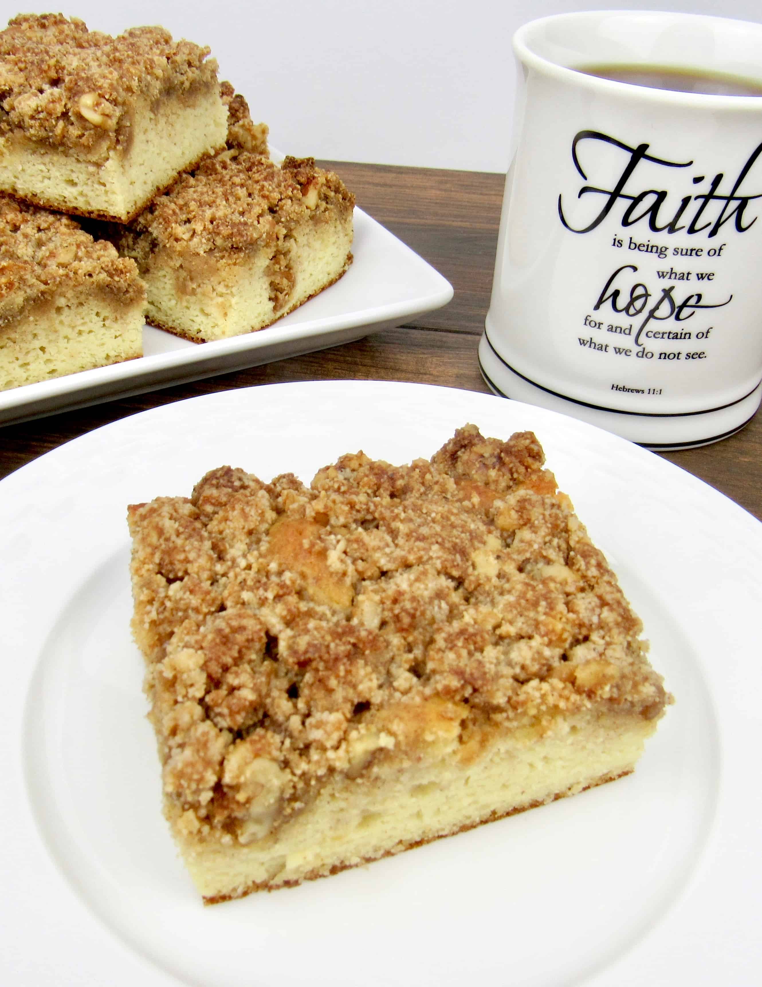 slice of coffee cake on white plate, cup of coffee and crumb cake slices in background