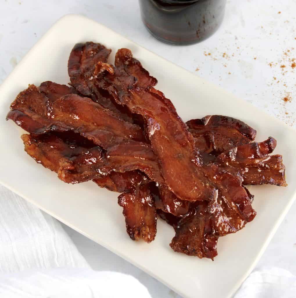 candied bacon stacked up on white plate with maple syrup in background