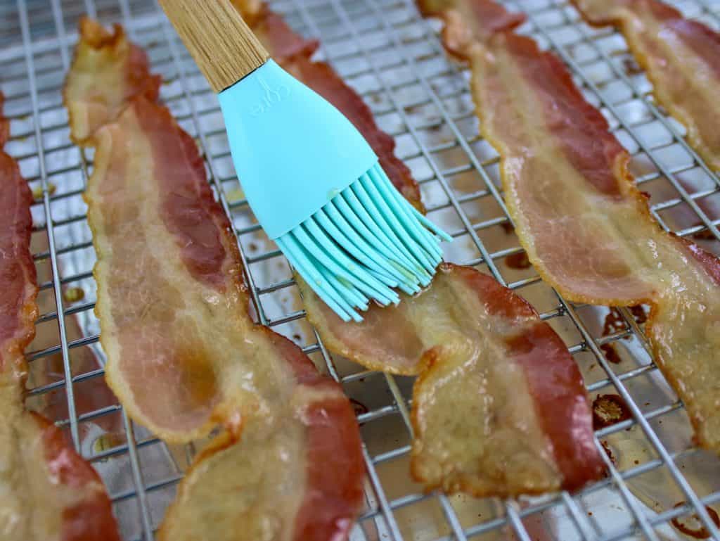 closeup of bacon on baking sheet being brushed with maple syrup