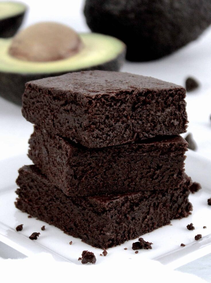 chocolate avocado brownies stacked up on white plate avocado in background