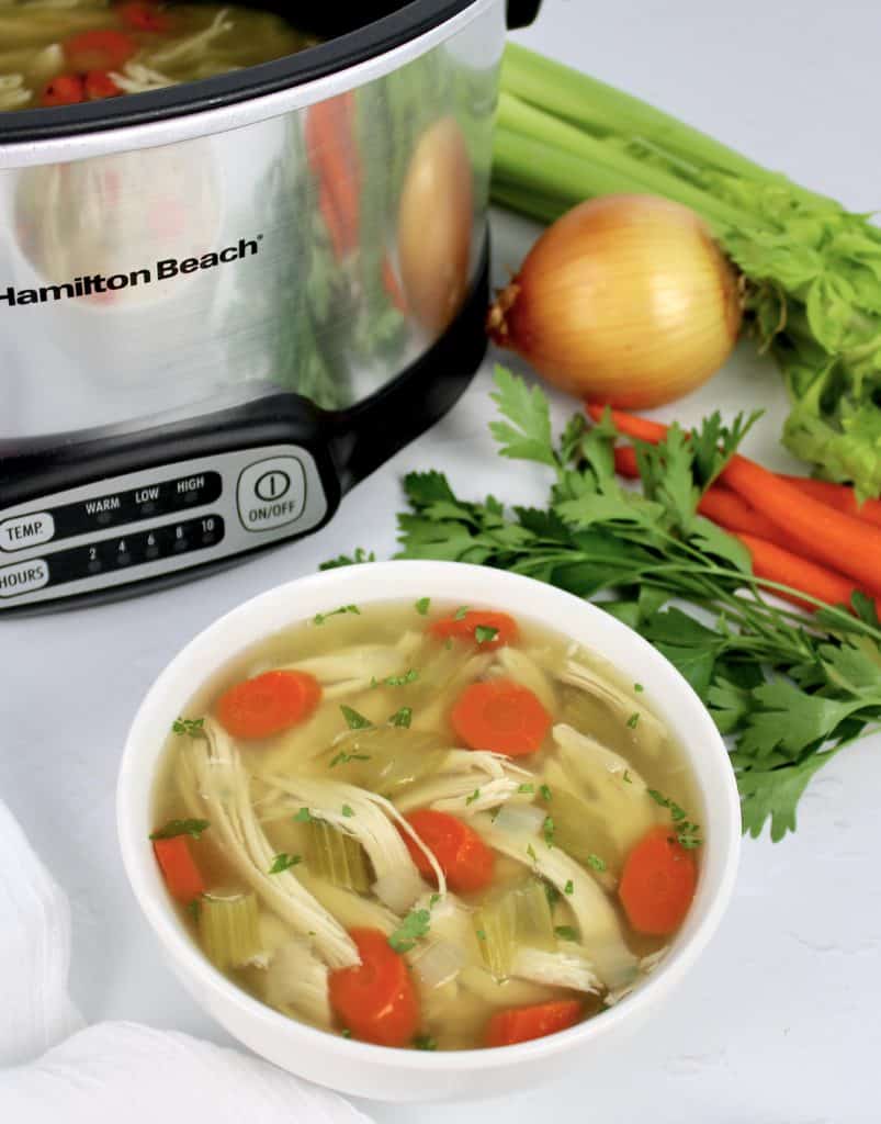 chicken soup in white bowl with veggies and crockpot in background