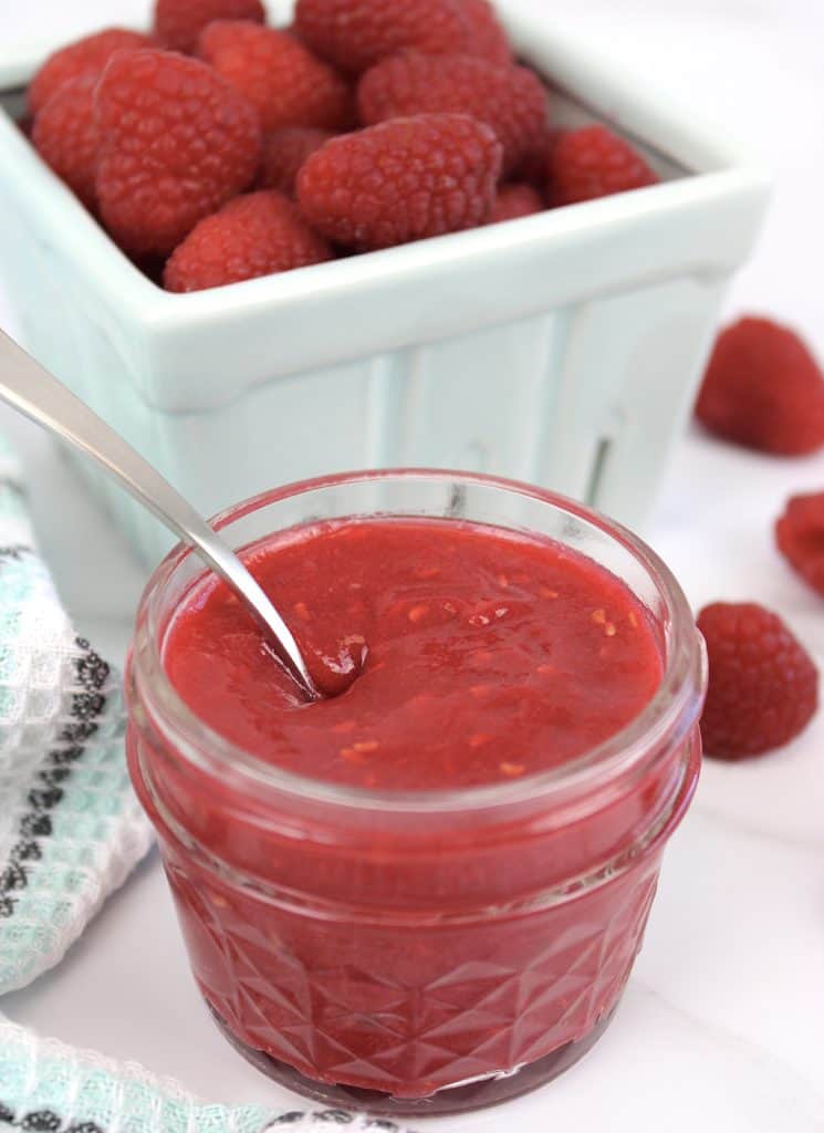 raspberry jam in jar with spoon and raspberries in background