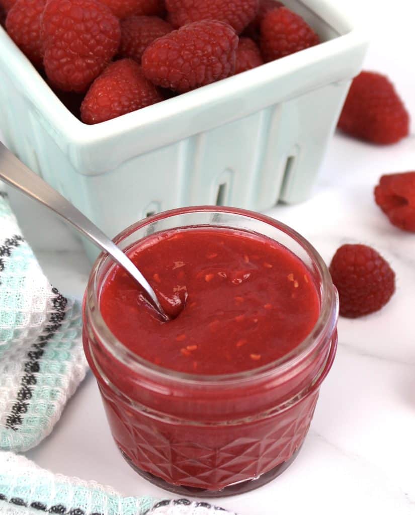 raspberry jam in jar with spoon and raspberries in background