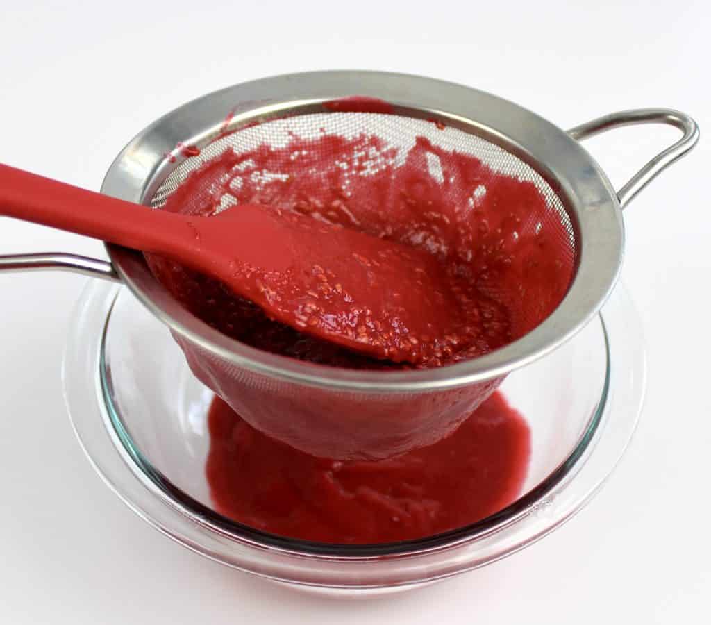 raspberry jam being strained with sieve in glass bowl