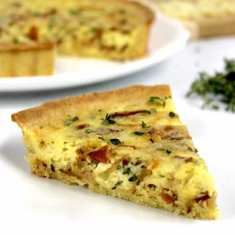 closeup of slice of quiche lorraine on white plate with rest of quiche in background