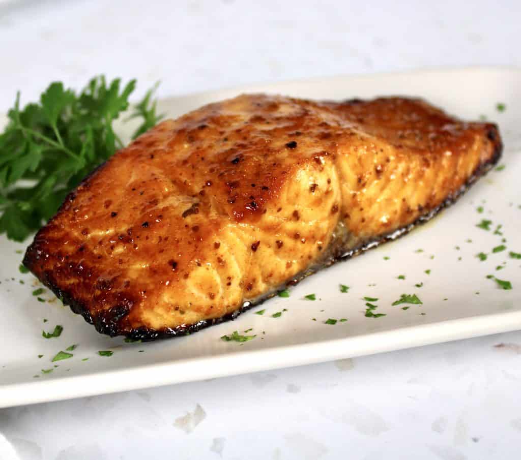piece of cooked glazed salmon on white plate with minced parsley around the plate
