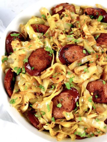 overhead view of fried cabbage with sausage in white bowl with parsley on top