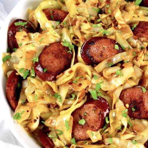 overhead view of fried cabbage with sausage in white bowl with parsley on top
