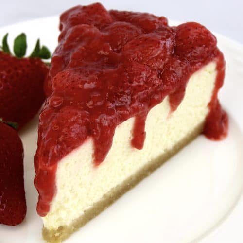 slice of cheesecake on white plate with strawberry sauce on top and strawberry on the side