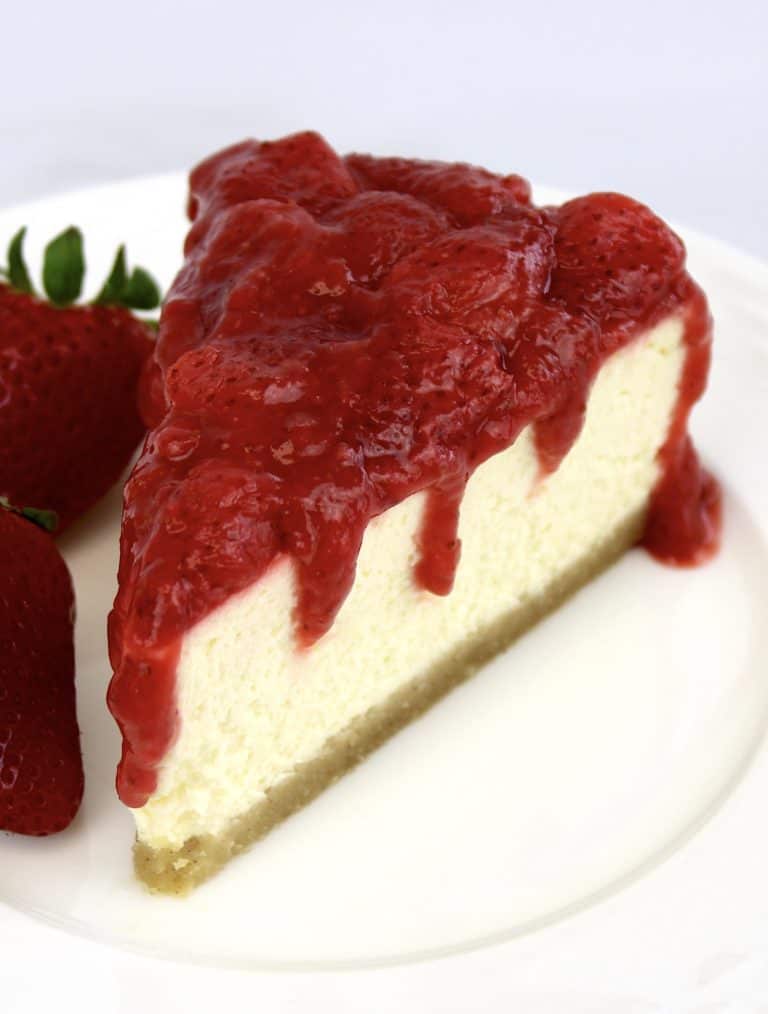 slice of cheesecake on white plate with strawberry sauce on top and strawberry on the side