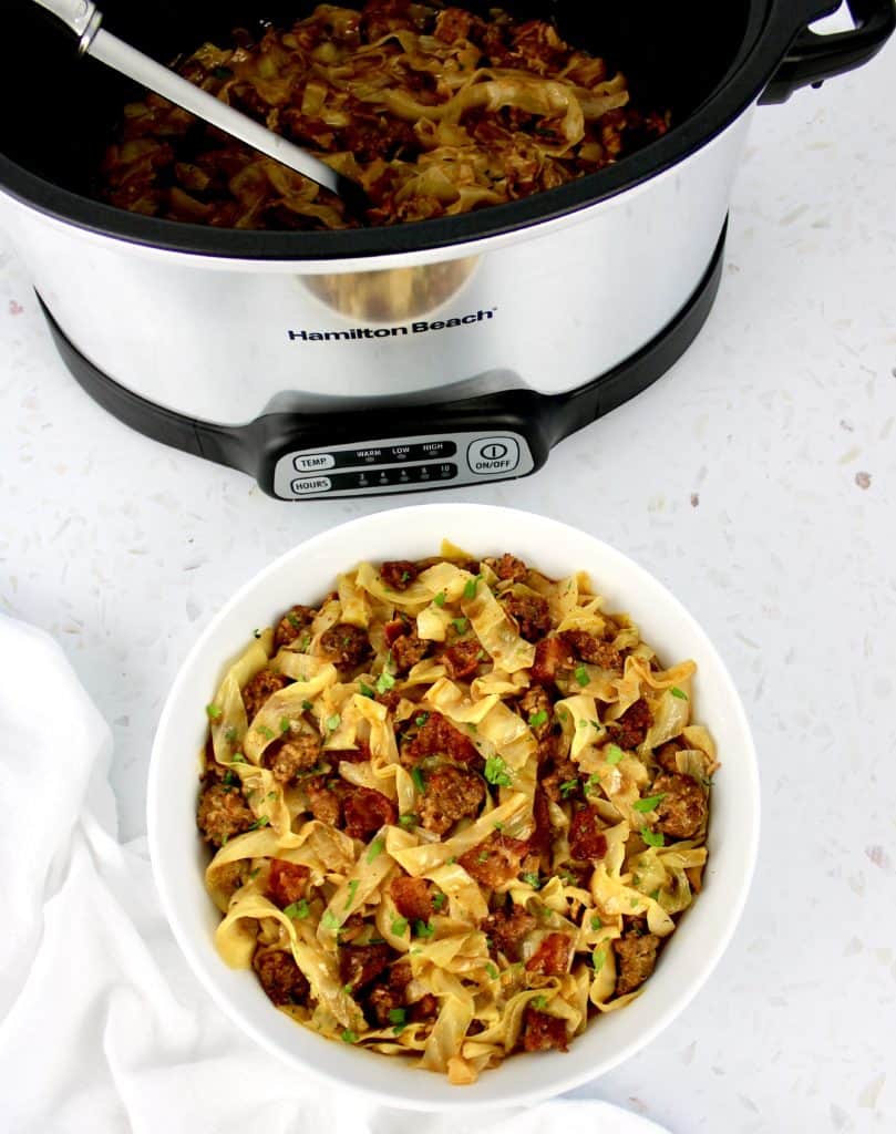 cabbage with sausage and bacon in white bowl with slow cooker in background