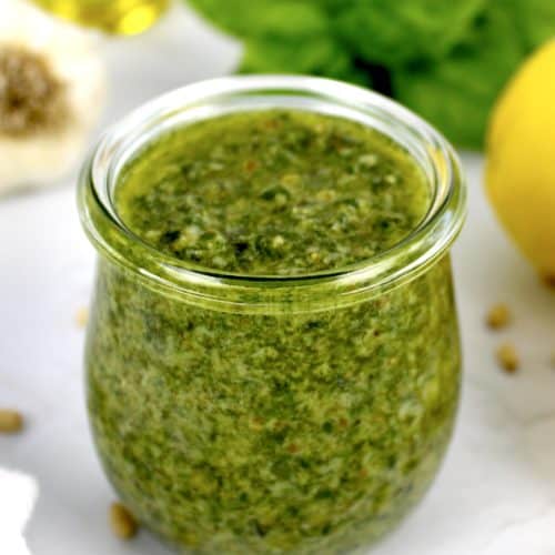 closeup of pesto sauce in open glass jar with lemons and basil in background
