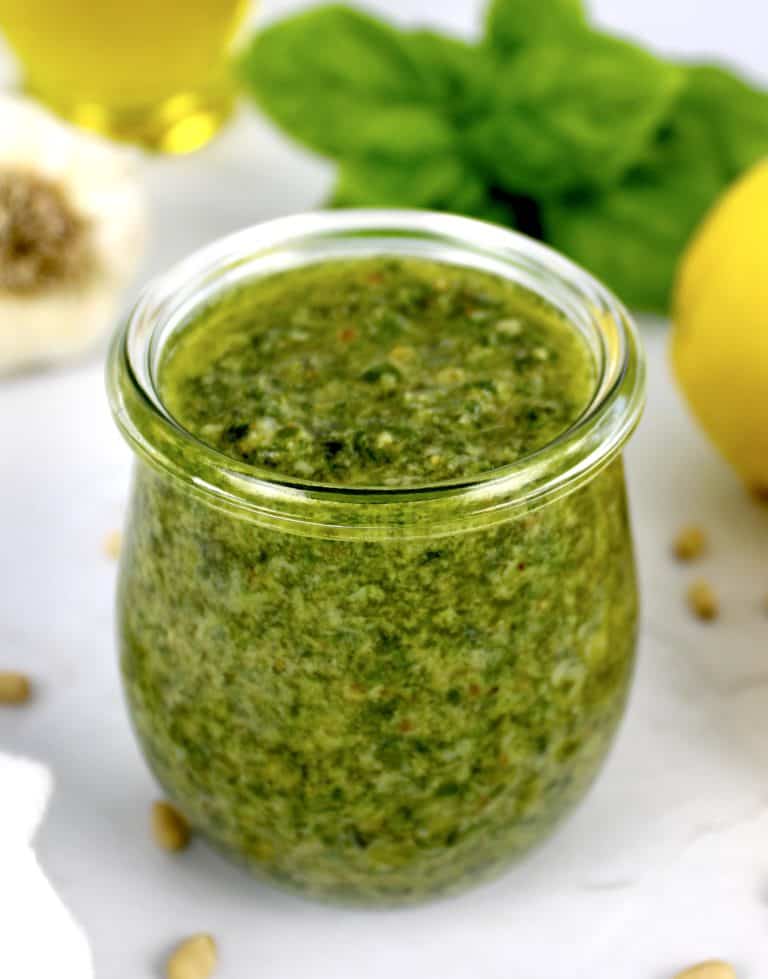closeup of pesto sauce in open glass jar with lemons and basil in background
