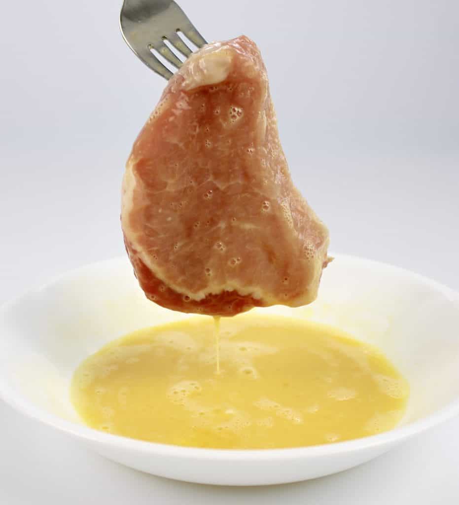 pork chop being dipped into egg in white bowl