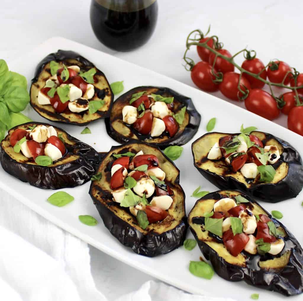 6 slices of grilled eggplant caprese with tomatoes in background