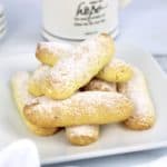 lady fingers stacked up on white plate with powdered sugar on top and coffee in background