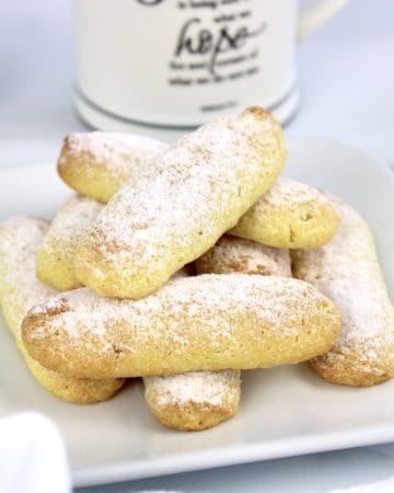 lady fingers stacked up on white plate with powdered sugar on top and coffee in background