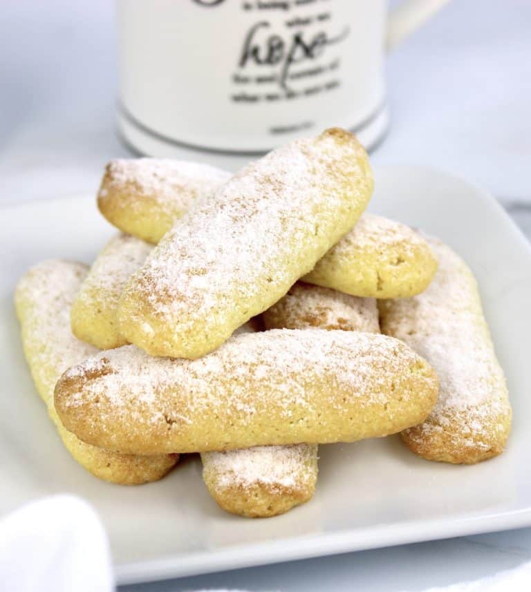 lady fingers stacked up on white plate with powdered sugar on top and coffee in background