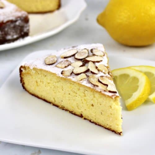 slice of lemon ricotta cake on white plate with cake in background
