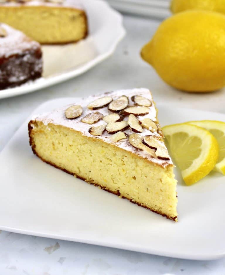 slice of lemon ricotta cake on white plate with cake in background