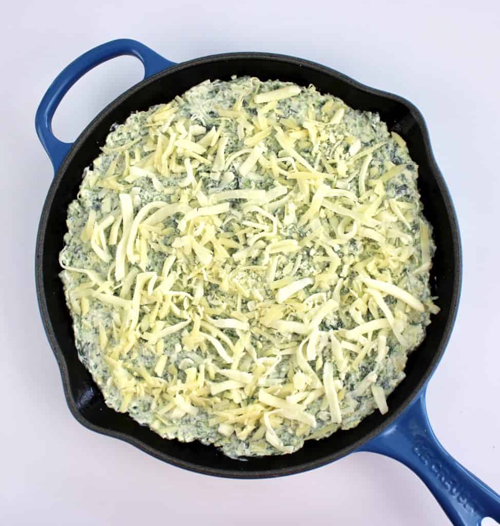 spinach artichoke dip mixture in cast iron skillet with shredded white cheese on top unbaked