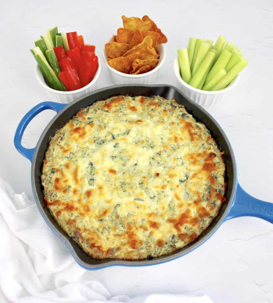 Spinach Artichoke Dip in blue pan with veggie sticks and tortilla chips in white bowls