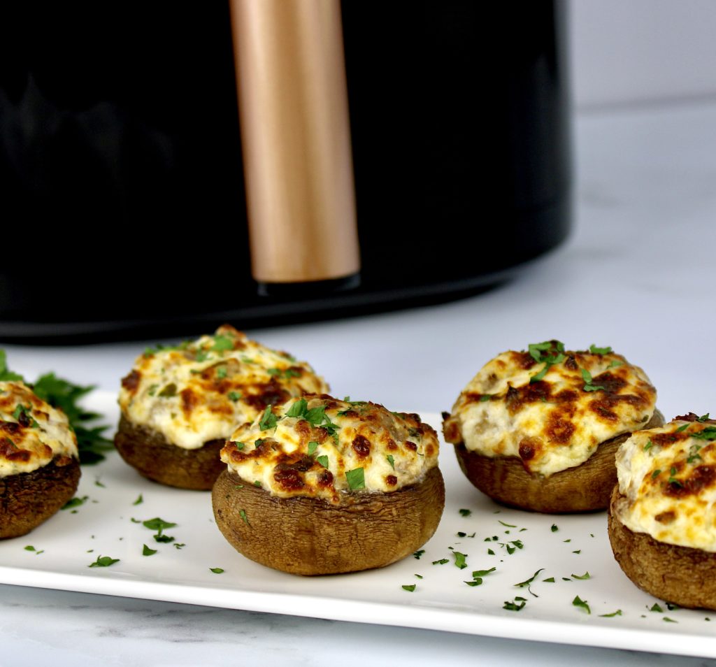 5 stuffed mushrooms on white plate with parsley garnish and air fryer in background