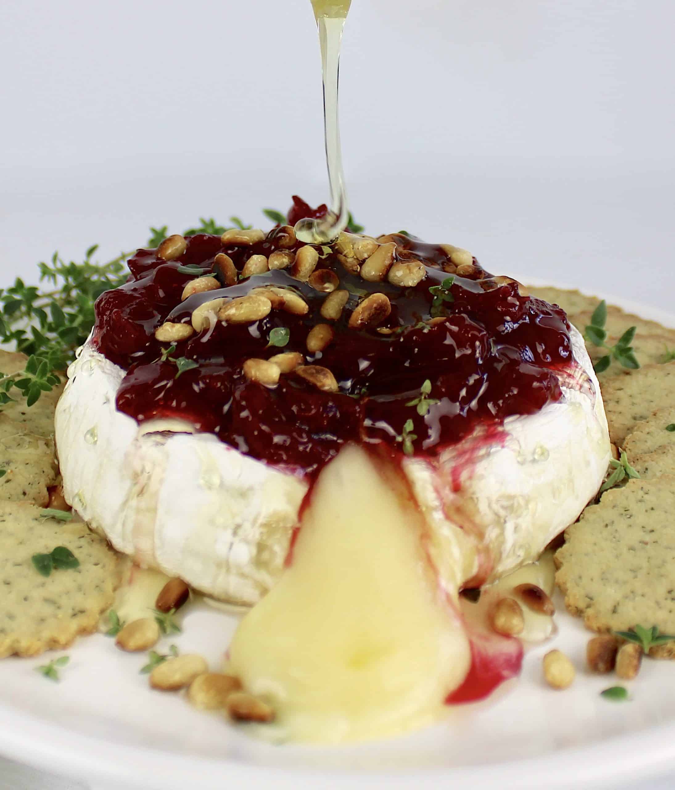 honey being drizzled over baked brie with cranberry sauce and toasted pine nuts