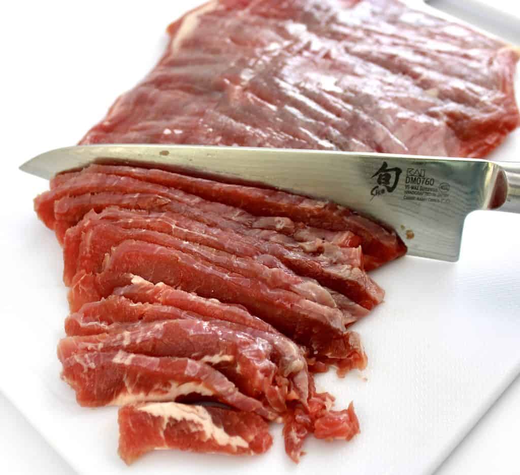 flank steak on white cutting board being sliced thinly with knife