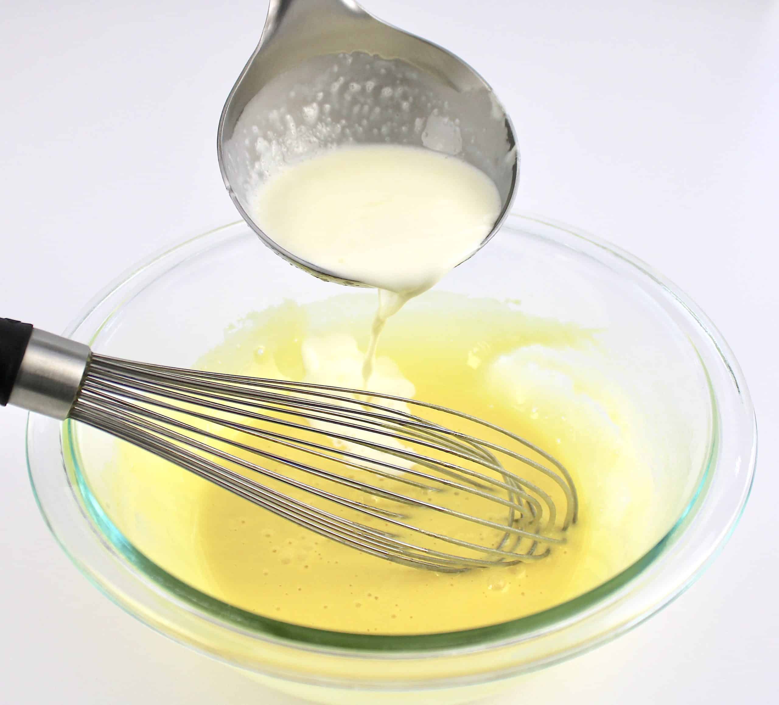cream being ladled into egg mixture in glass bowl with whisk