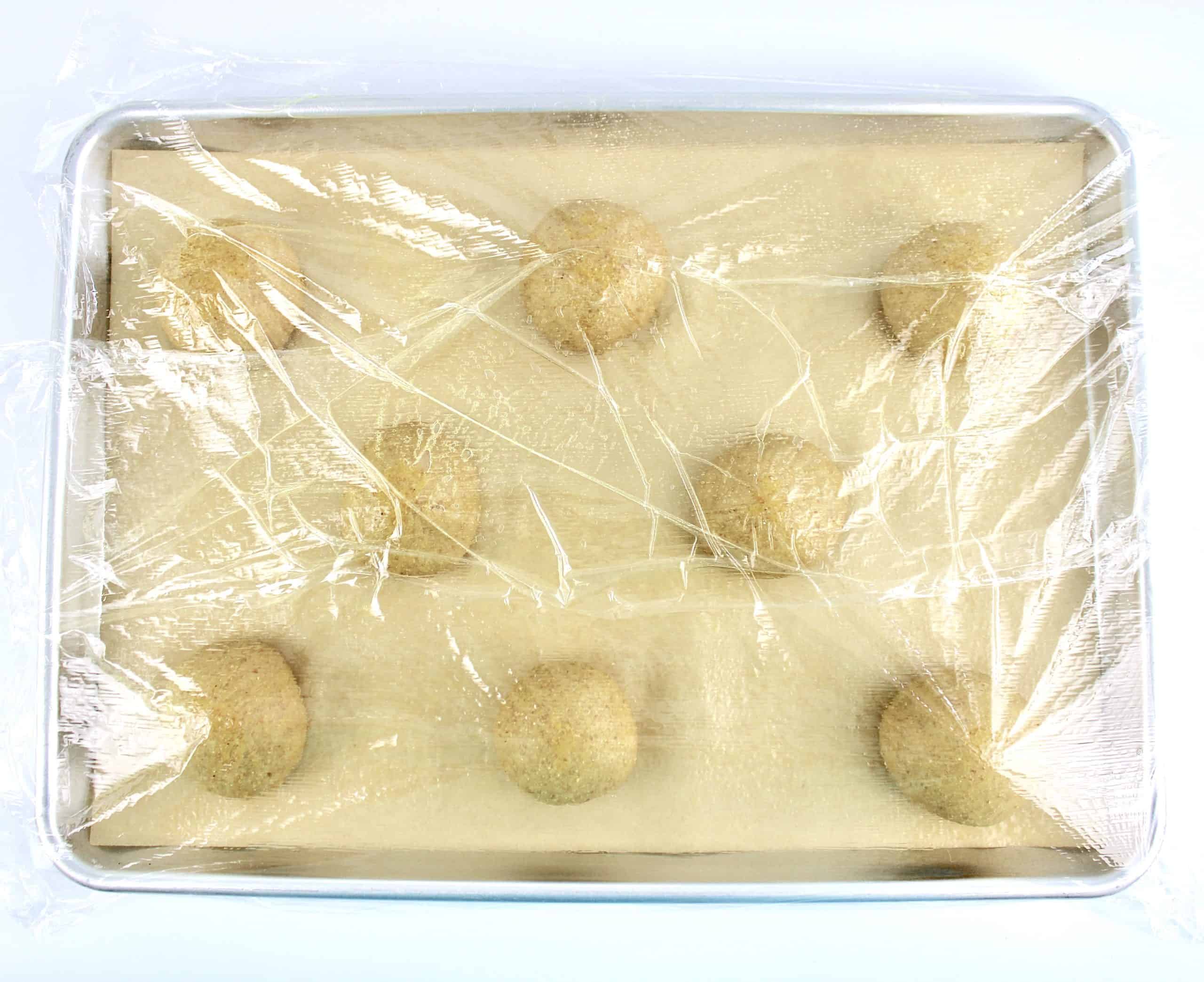 8 dough balls in baking sheet with parchment and plastic wrap on top