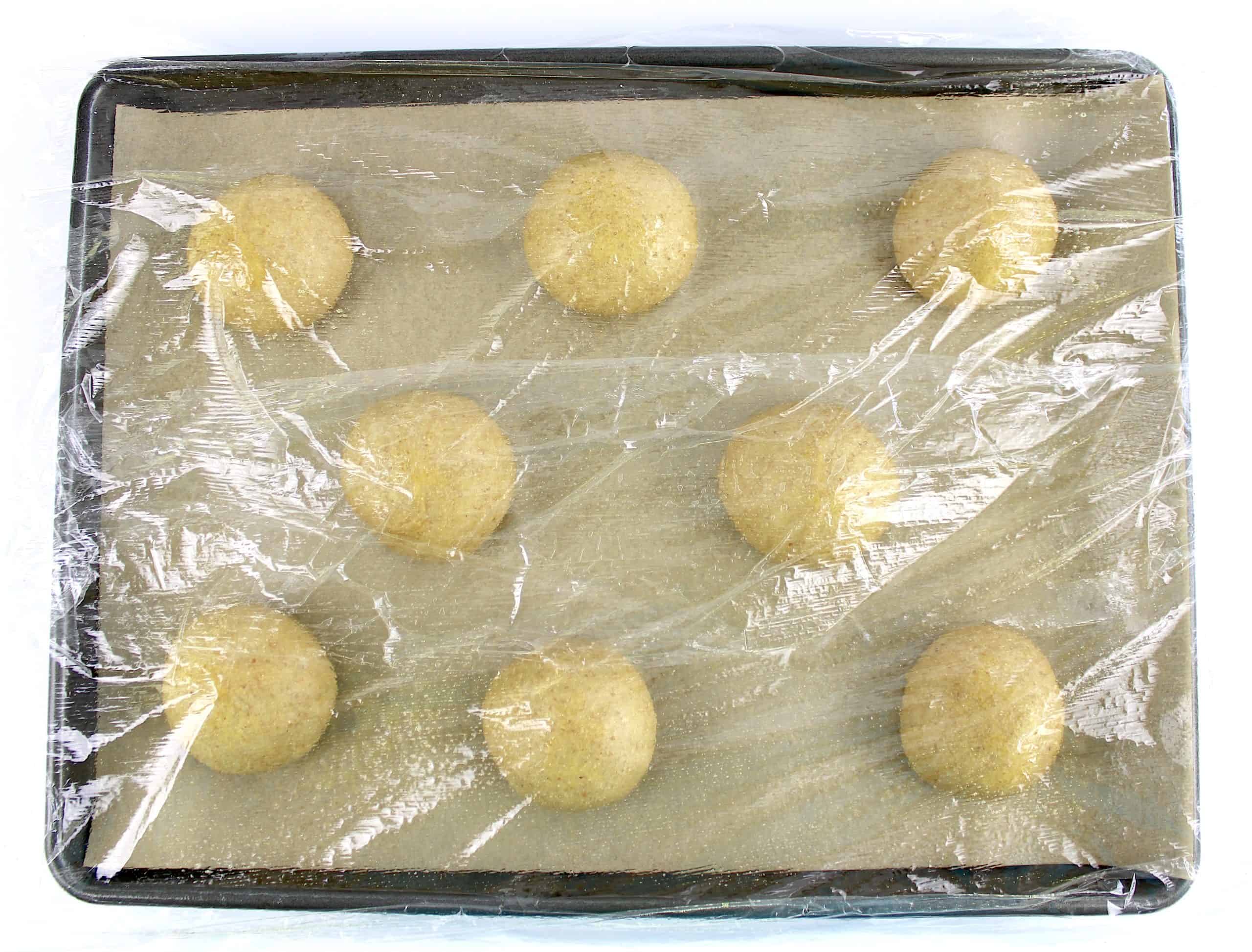 8 keto bun dough balls on parchment lined baking sheet with plastic wrap on top