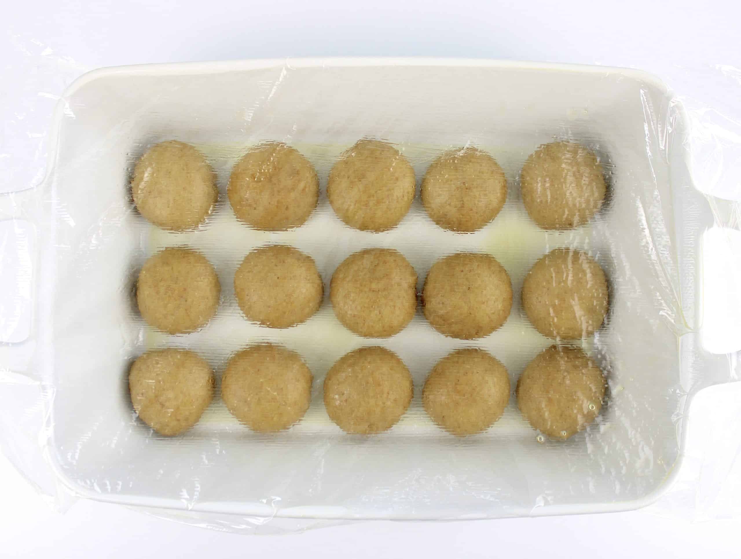 15 dinner rolls in white baking dish with plastic wrap on top