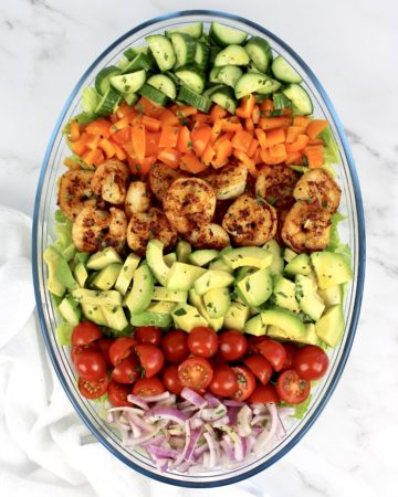 salad with rows of shrimp avocado tomatoes peppers onions in glass bowl
