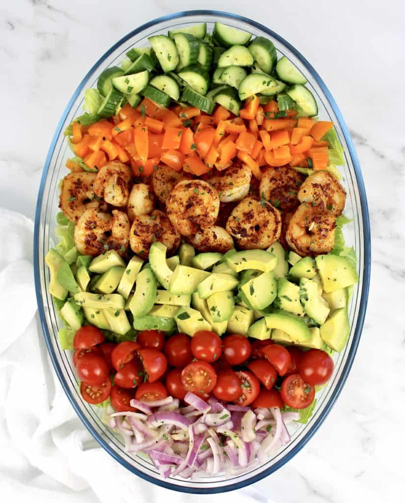 salad with rows of shrimp avocado tomatoes peppers onions in glass bowl