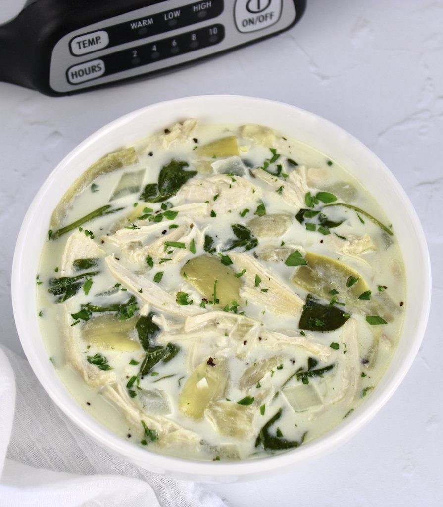Spinach Artichoke Chicken Soup in white bowl with slow cooker in background