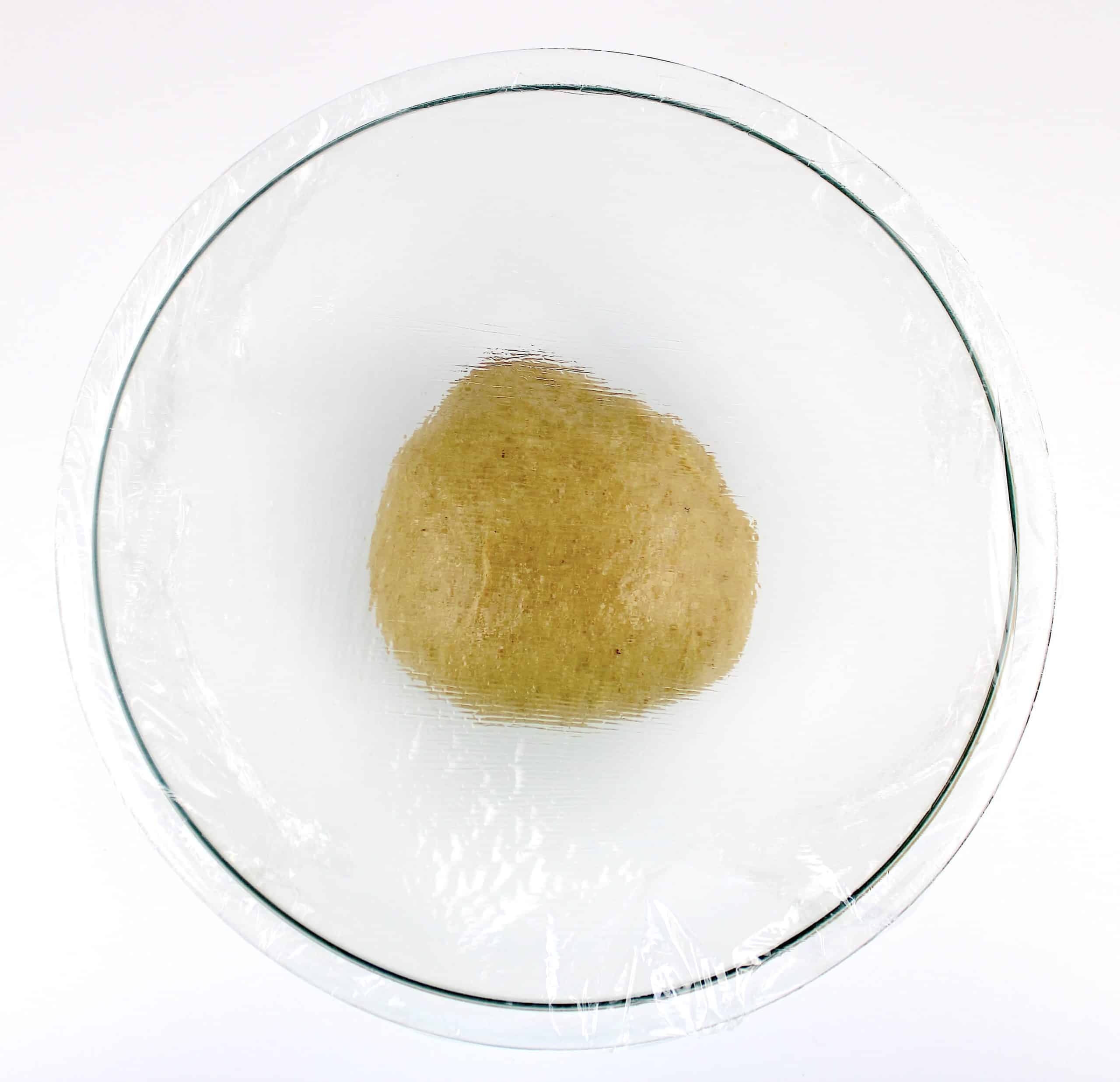 cinnamon roll dough ball in glass bowl with plastic wrap on top