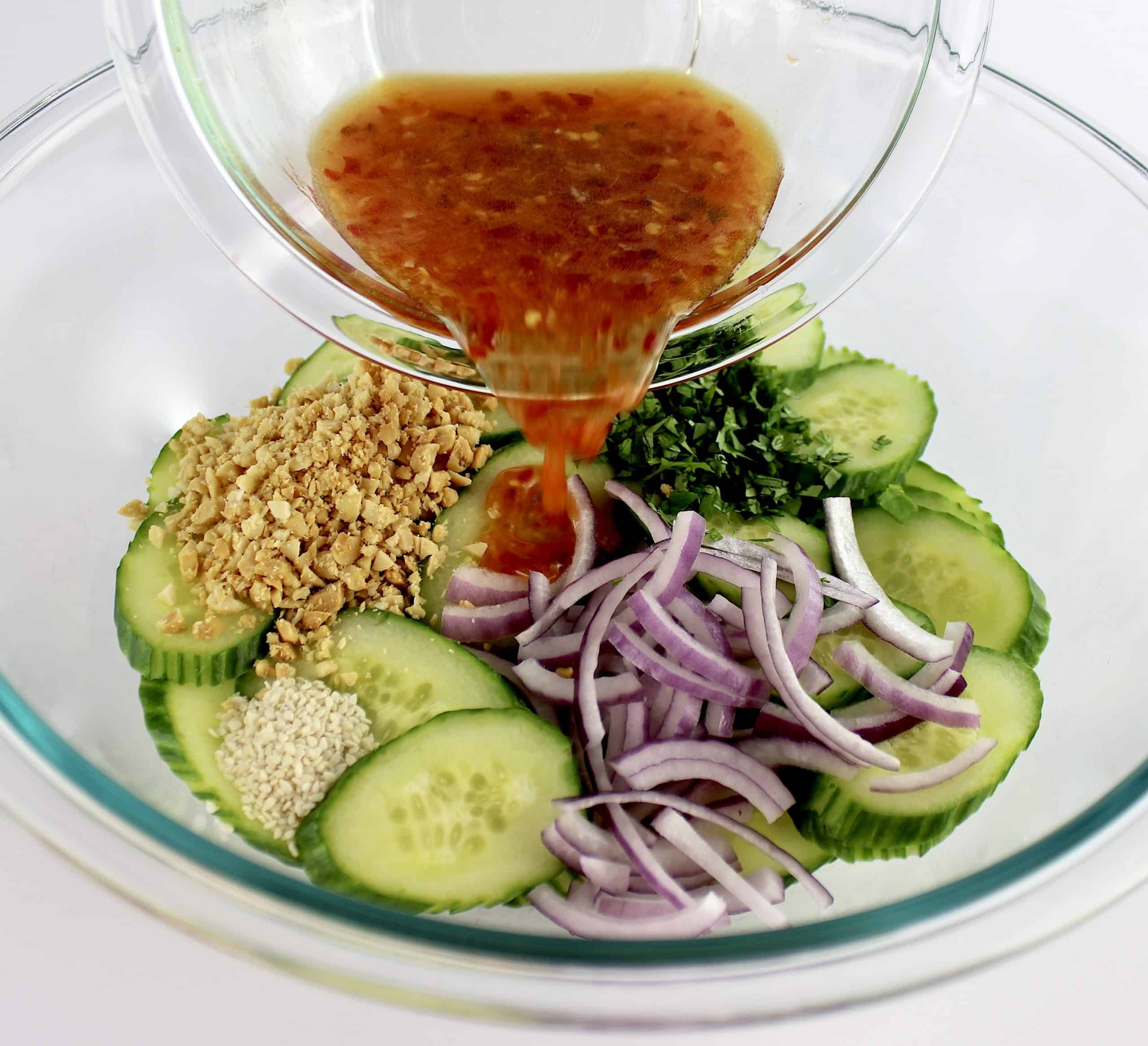 dressing being poured over cucumber salad in glass bowl