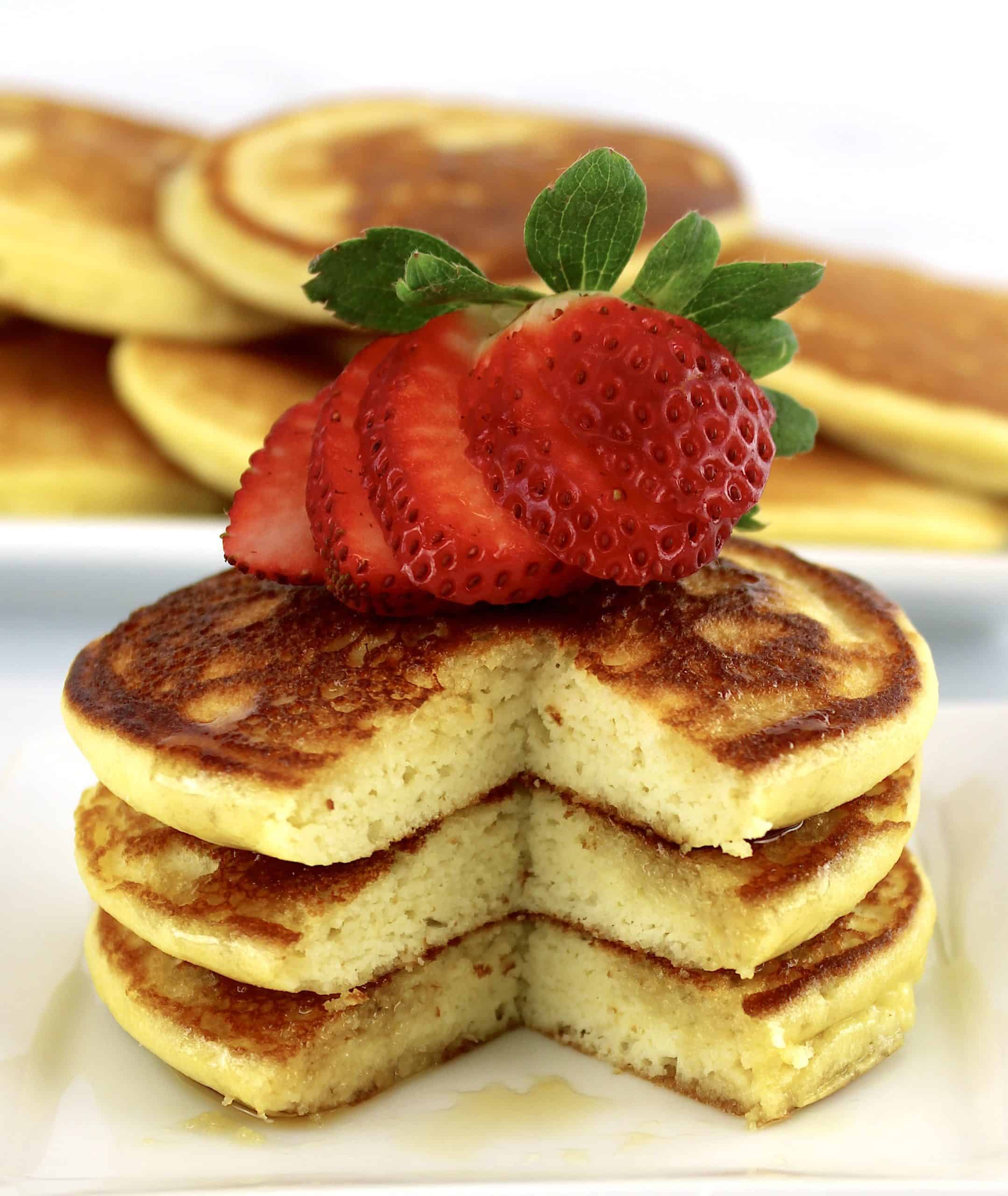 3 pancakes on white plate in stack with sliced strawberry on top and slice missing from pancakes