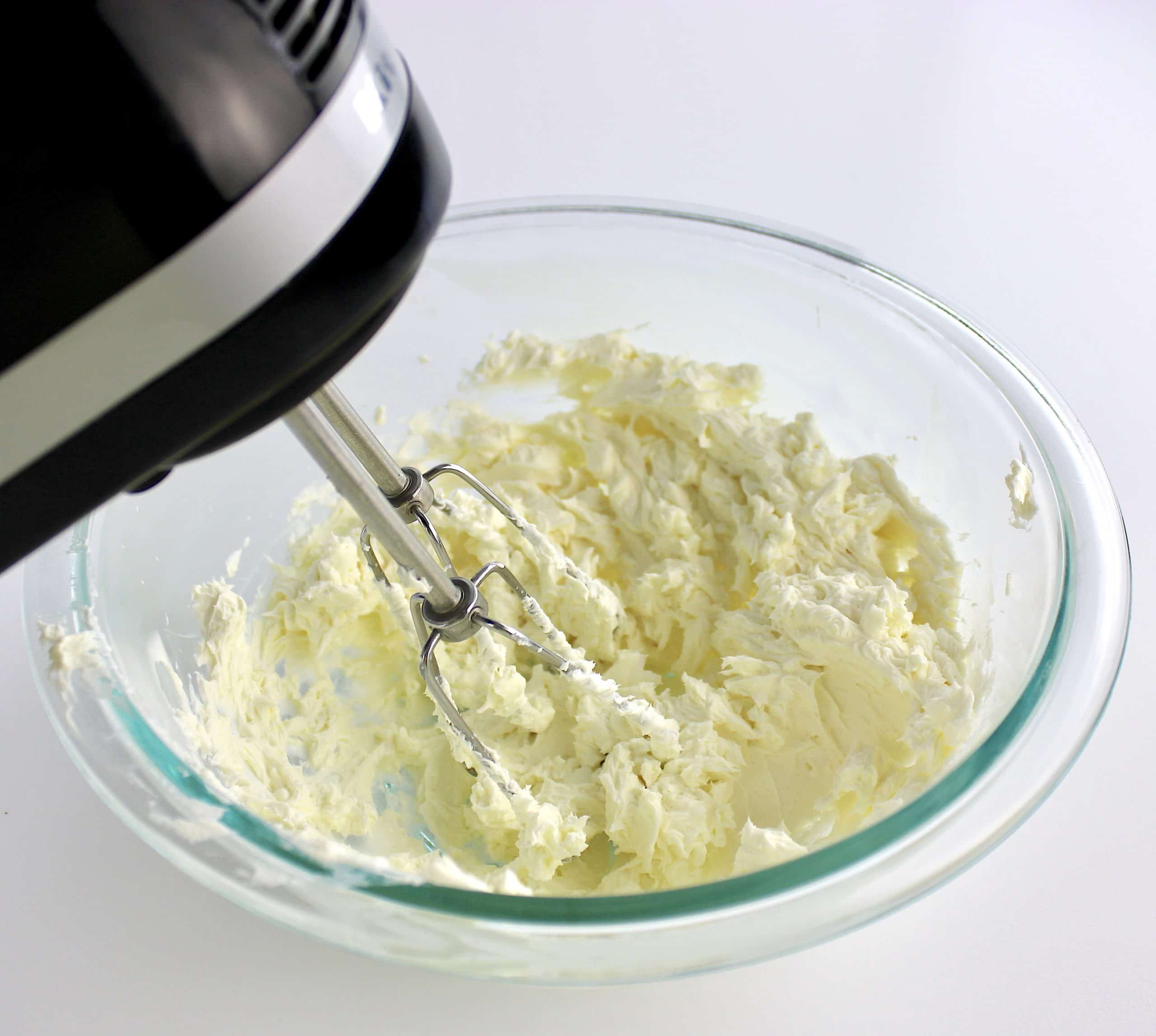 cream cheese being whipped with hand mixer in glass bowl