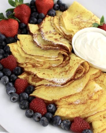Keto Crepes folded on platter with whip cream in bowl and berries all around