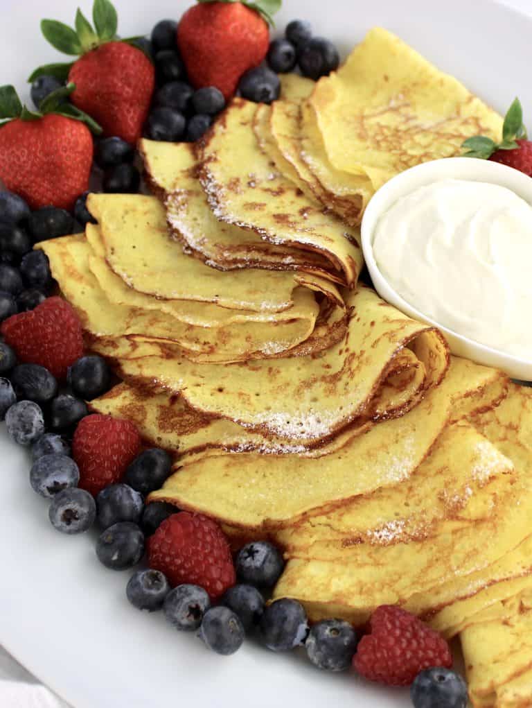 Keto Crepes folded on platter with whip cream in bowl and berries all around
