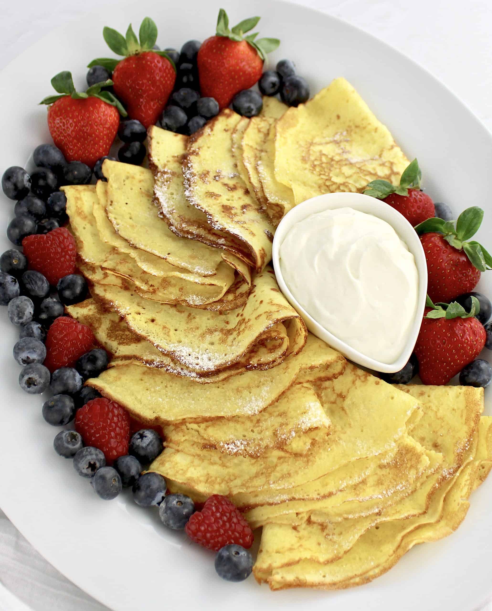 Keto Crepes on white platter with whip cream in bowl and berries all around