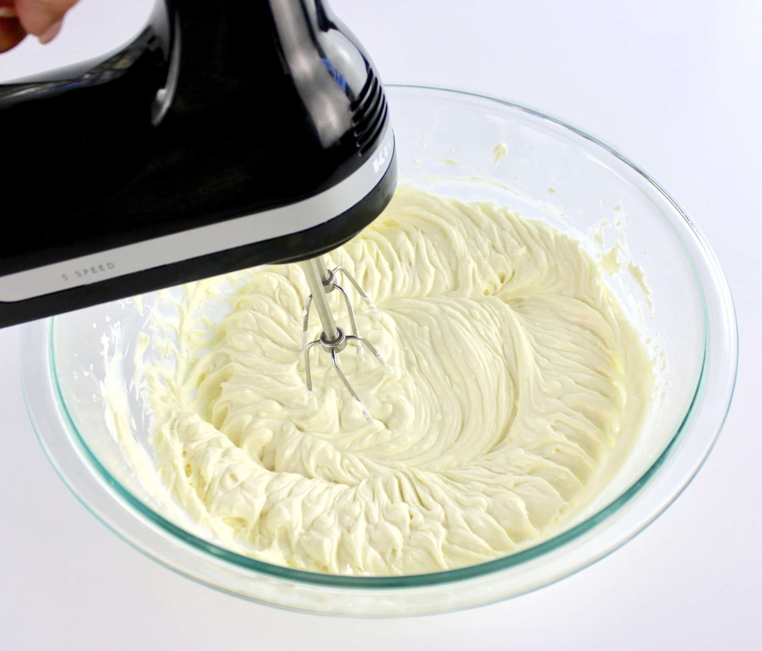 cheesecake filling being whipped with hand mixer in glass bowl