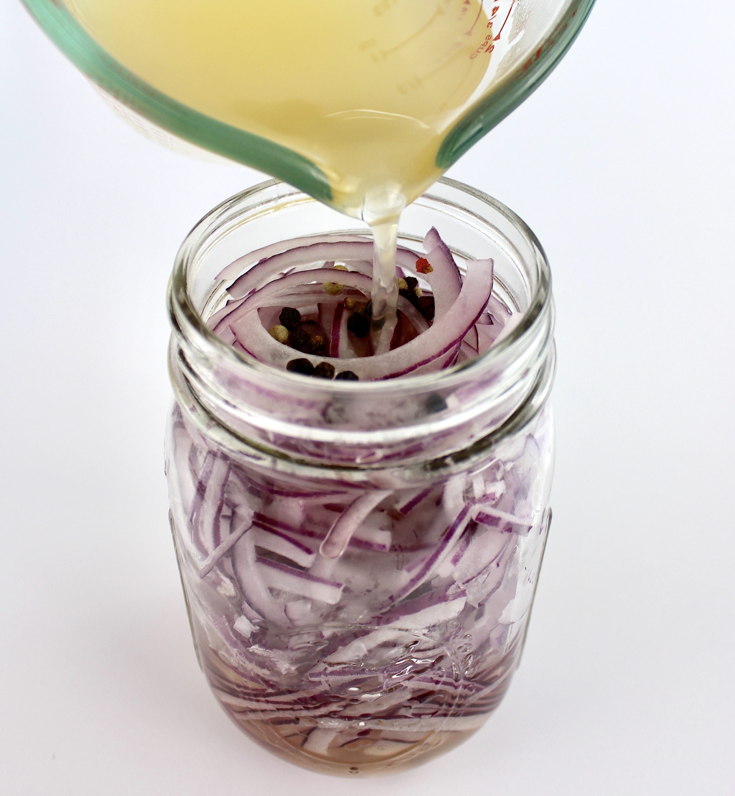 vinegar being poured over sliced onions and peppercorns in open mason jar