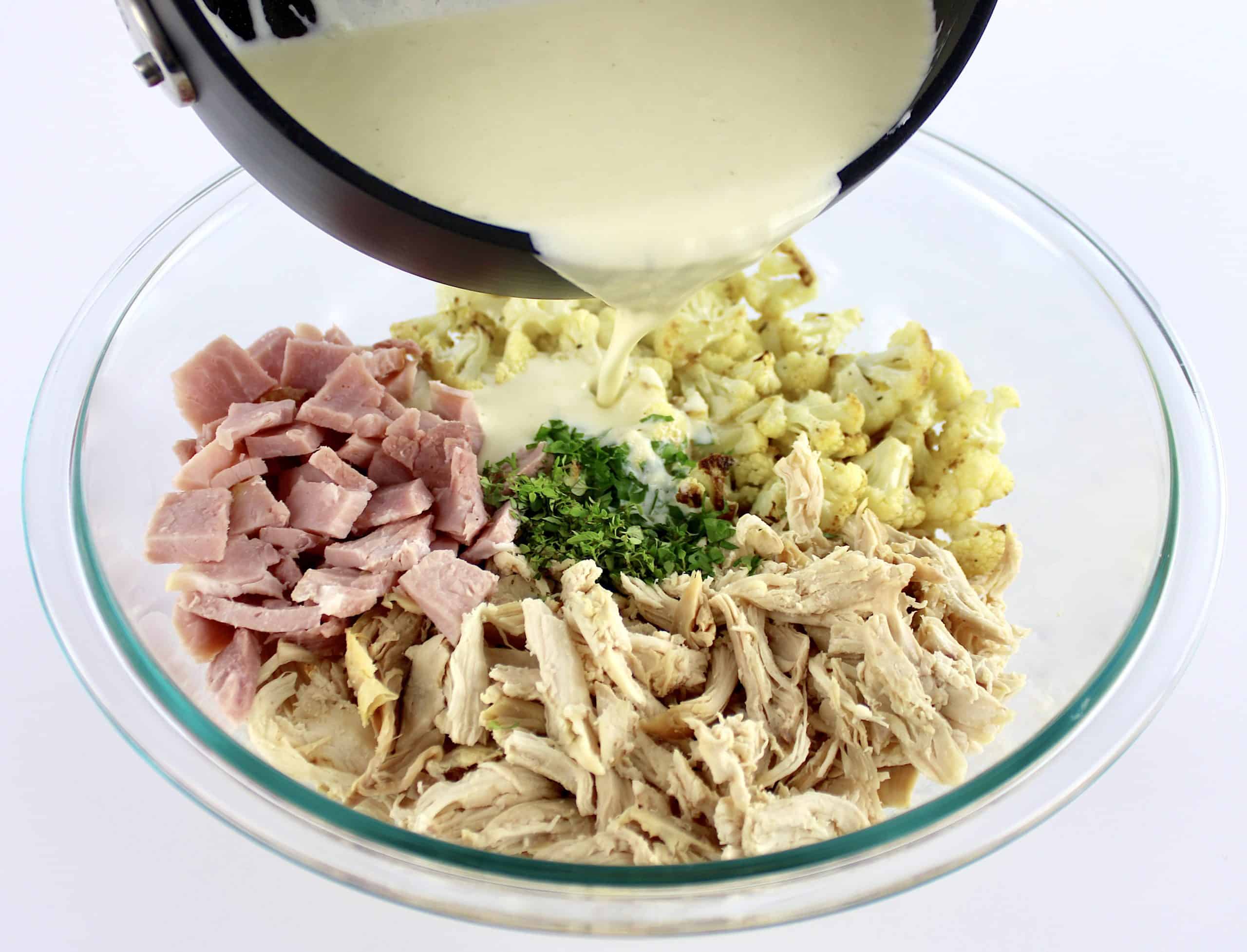 cheese sauce being poured over chicken ham and roasted cauliflower in glass bowl